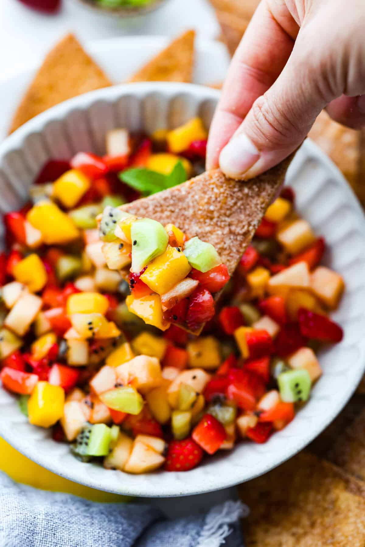 Vibrant fruit salsa is a fresh, juicy twist on your favorite snack! Sweet berries, tropical mango, and a splash of lime always steal the spotlight! Especially when served with crispy cinnamon chips! Someone taking a scoop of fruit salsa with a cinnamon sugar chip.