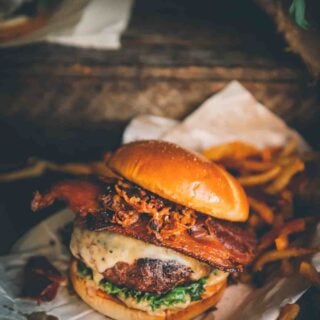 Huge ground venison burger on a soft bun with lettuce, cheese melting down the side, thick cut bacon, and crispy shallots with fries in the background.
