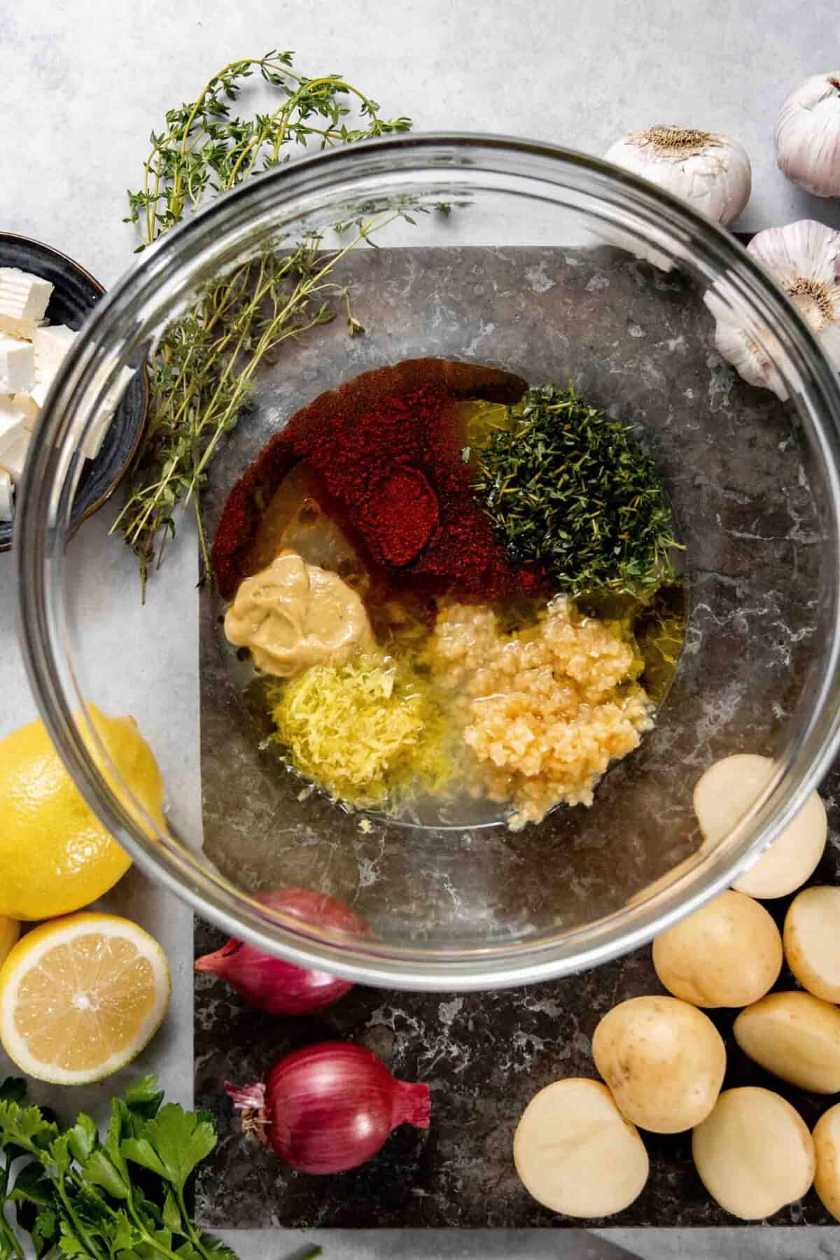 A glass bowl containing various ingredients such as chopped herbs, spices, garlic, mustard, and lemon zest, placed on a marble cutting board surrounded by garlic heads, onions, lemons, and baby potatoes.