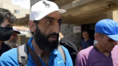 BBC / Jon Donnison Kiness Abu Motab wears a white ralph lauren designer baseball cap, a grey backpack with straps and a blue slazenger polo shirt. He is looking away from the camera at a reporter out of shot