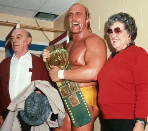 Getty Images Hulk, with an open-mouthed smile, covered in sweat and holding a gold belt, standing between his parents backstage at Madison Square Garden in New York in 1984