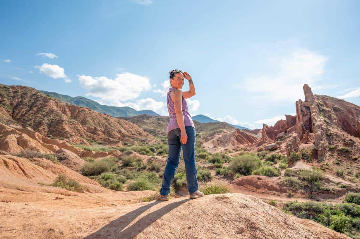 A woman stands on a rocky outcrop in Fairytale Canyon, Kyrgyzstan, with a backdrop of colorful, eroded sandstone cliffs and green vegetation under a bright, clear sky. She is wearing a light purple tank top and blue jeans, looking back over her shoulder with a smile, capturing the beauty and serenity of the canyon.
