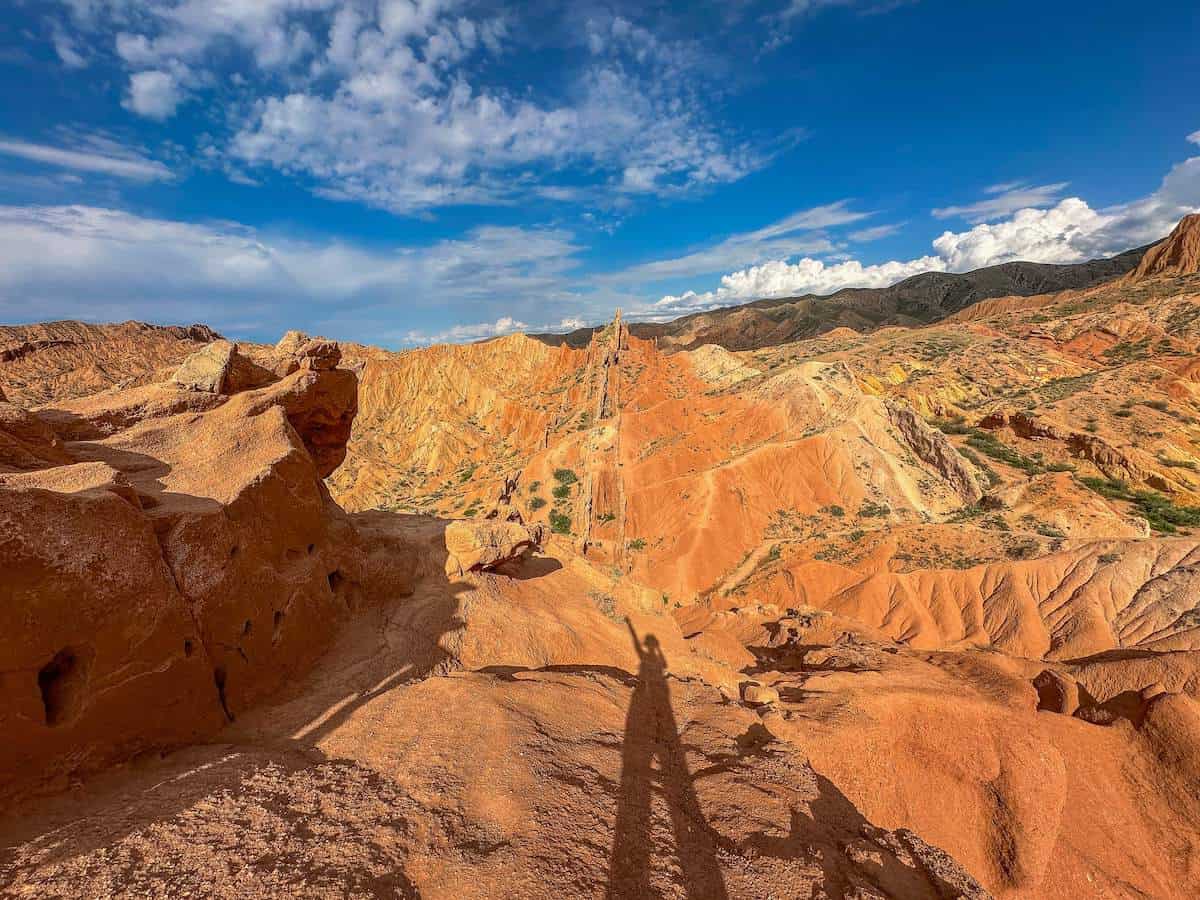 The Chinese Wall rock formation in Fairytale Canyon, Kyrgyzstan, with its dramatic, jagged, reddish-orange cliffs extending into the distance under a bright blue sky. The sunlight casts deep shadows, emphasizing the rugged terrain and highlighting the intricate erosion patterns that give the formation its distinctive appearance. This breathtaking view captures the unique geological features that make Fairytale Canyon a must-visit destination.